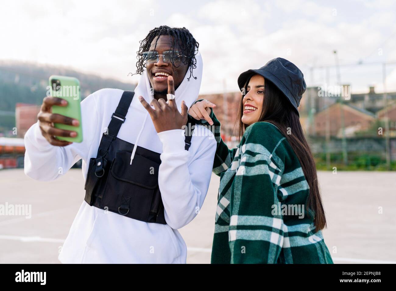 Young content multiracial partners in stylish apparel taking selfie on urban pavement in sunlight Stock Photo