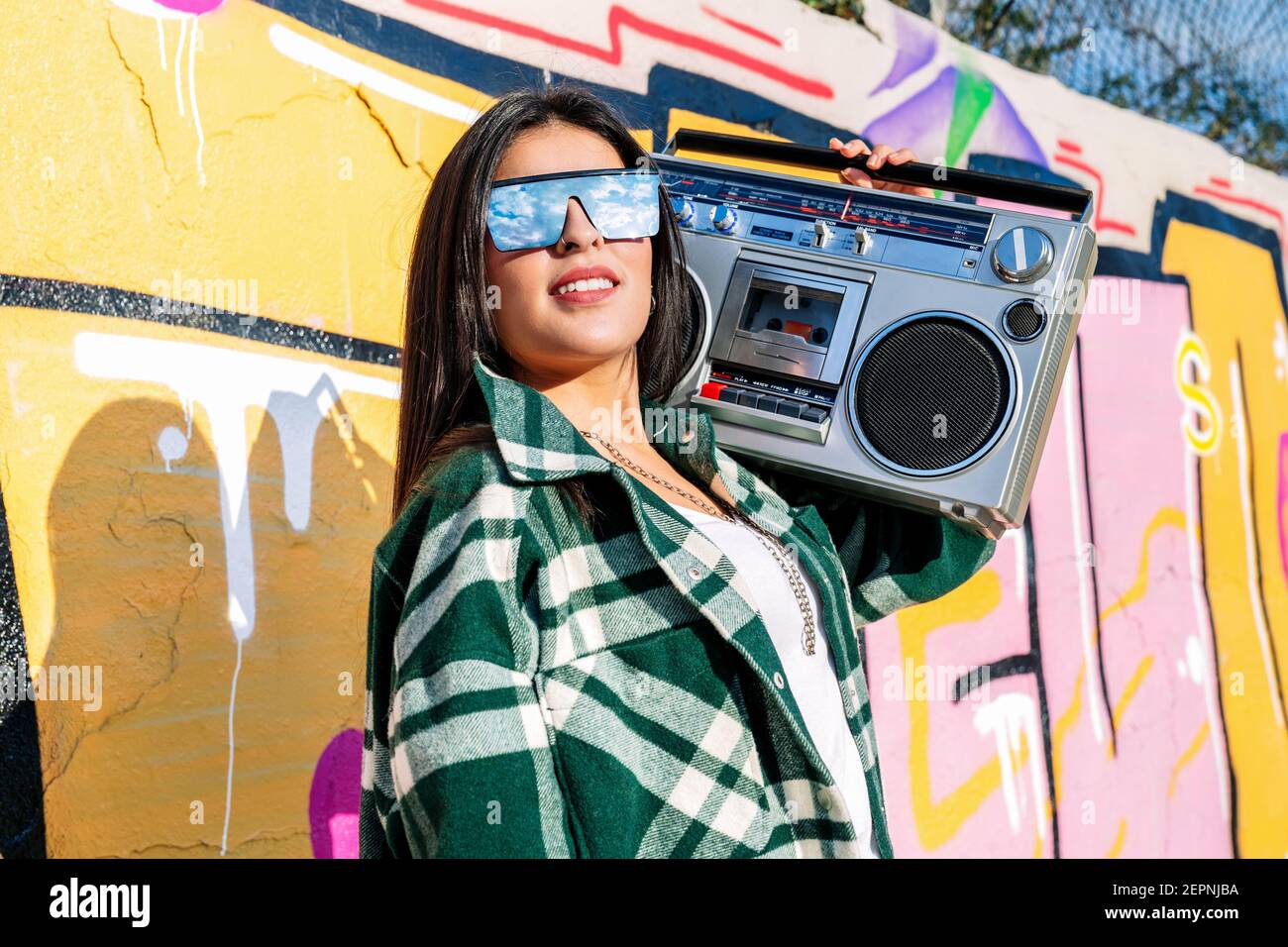 Young trendy female in sunglasses reflecting cloudy sky listening to song from old tape recorder against graffiti wall in sunlight Stock Photo