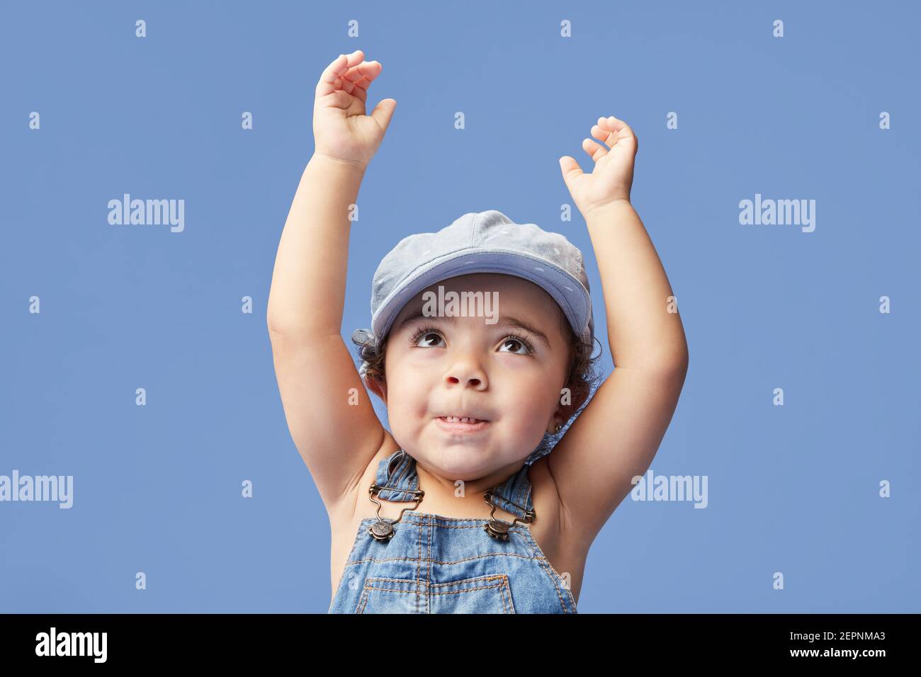 Charming barefoot child in denim dress and dress with curly hair looking up with arms raised while dancing on blue background Stock Photo