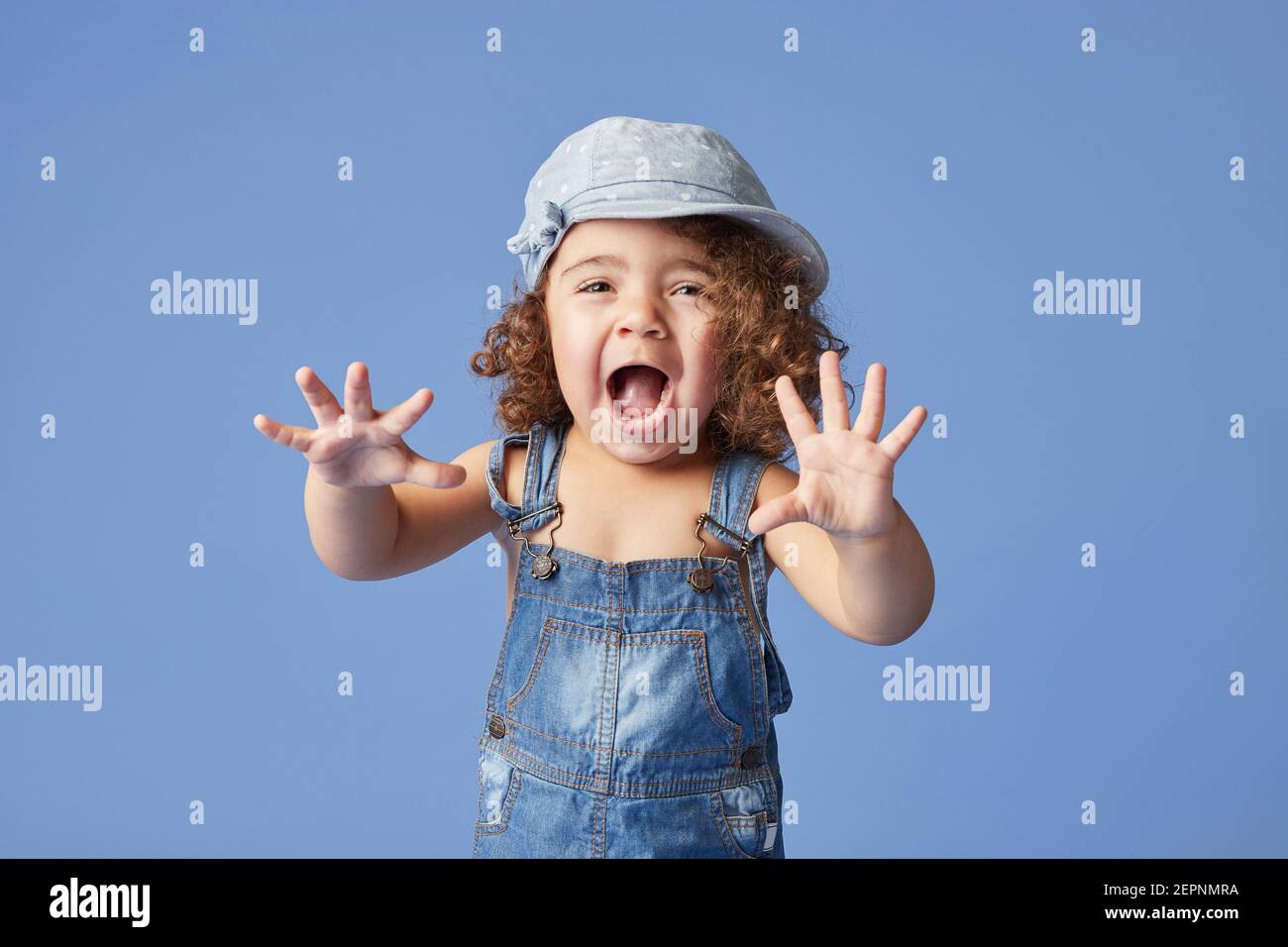 Charming barefoot child in denim dress and hat with curly hair looking a camera while standing on blue background making faces Stock Photo