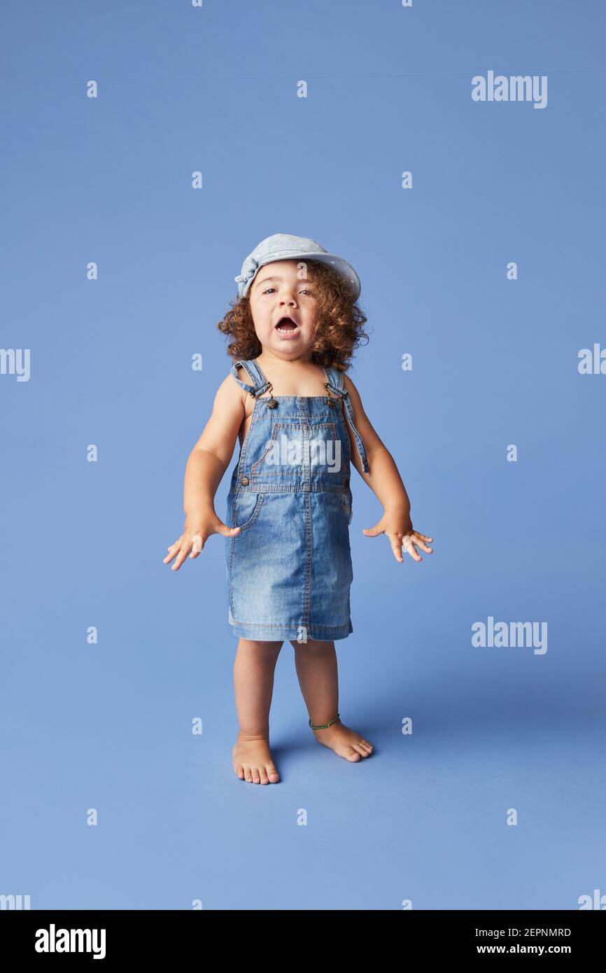 Charming cheerful barefoot child in denim dress and hat with curly hair looking at camera while dancing on blue background Stock Photo