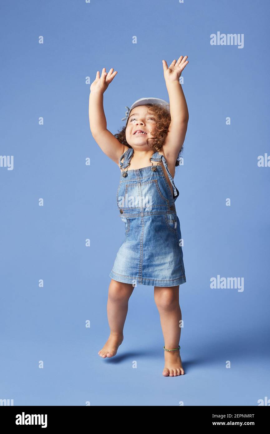 Charming barefoot child in denim dress and hat with curly hair looking up with arms raised while dancing on blue background Stock Photo