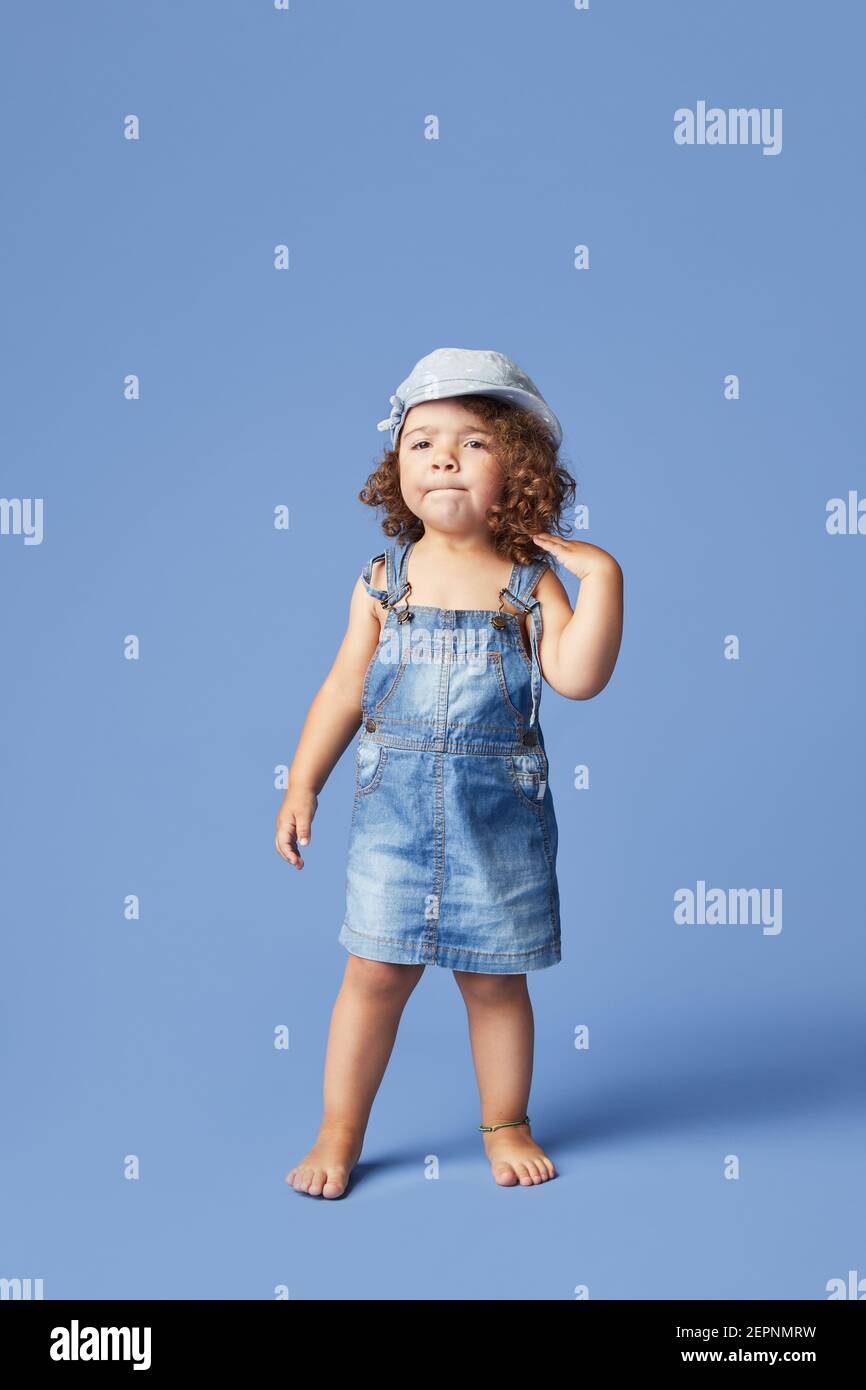 Charming barefoot child in denim dress and hat with curly hair looking away while dancing on blue background Stock Photo