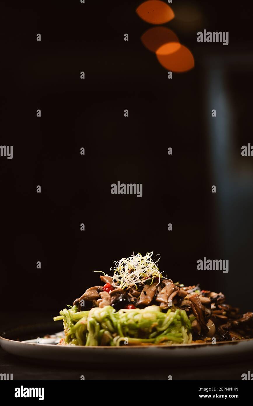 Low angle of yummy vegan dish with zucchini spaghetti and sauteed mushroom slices covered with red berries and alfalfa sprouts on dark background Stock Photo