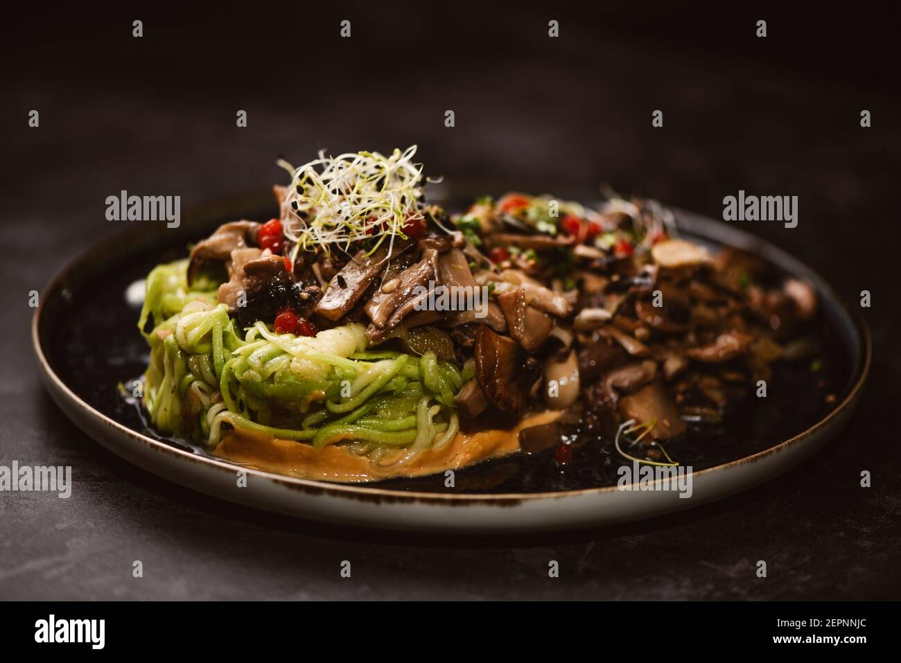 Yummy vegan dish with zucchini spaghetti and sauteed mushroom slices covered with red berries and alfalfa sprouts on dark background Stock Photo