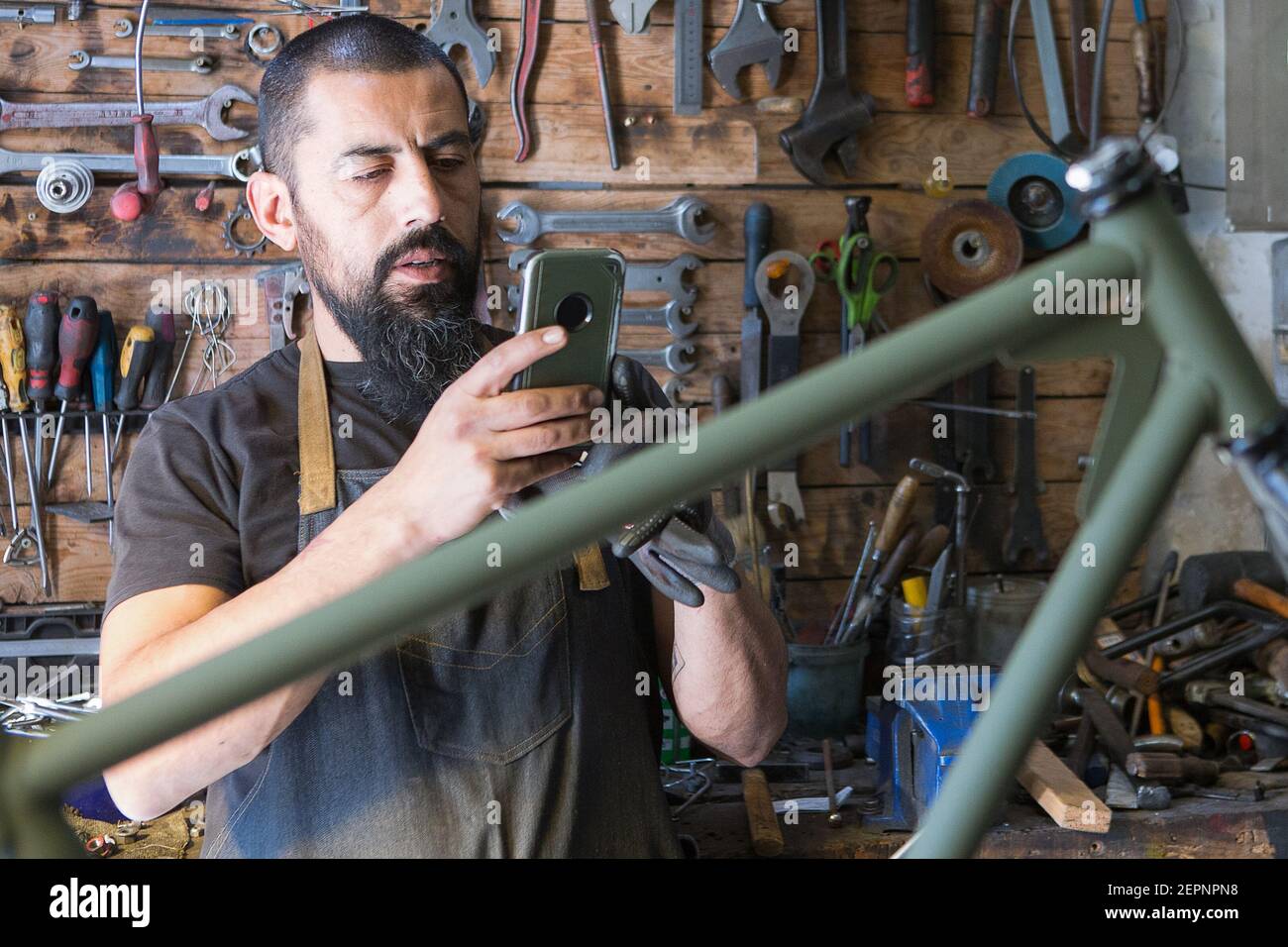 Concentrated bearded male master in apron taking photo of bicycle during repairing in garage Stock Photo