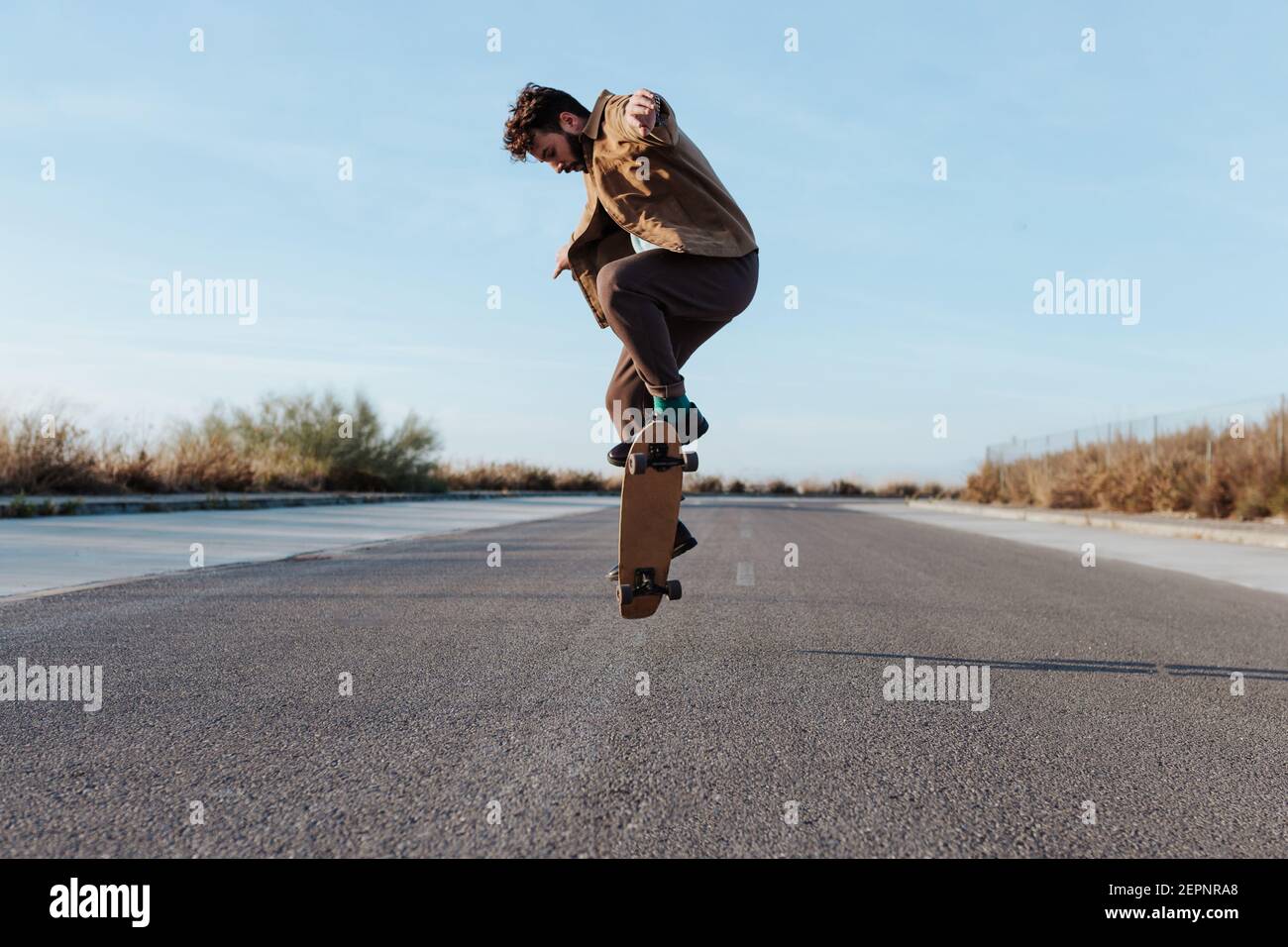 Full body young bearded skater in casual outfit jumping while performing kickflip on skateboard on asphalt road Stock Photo