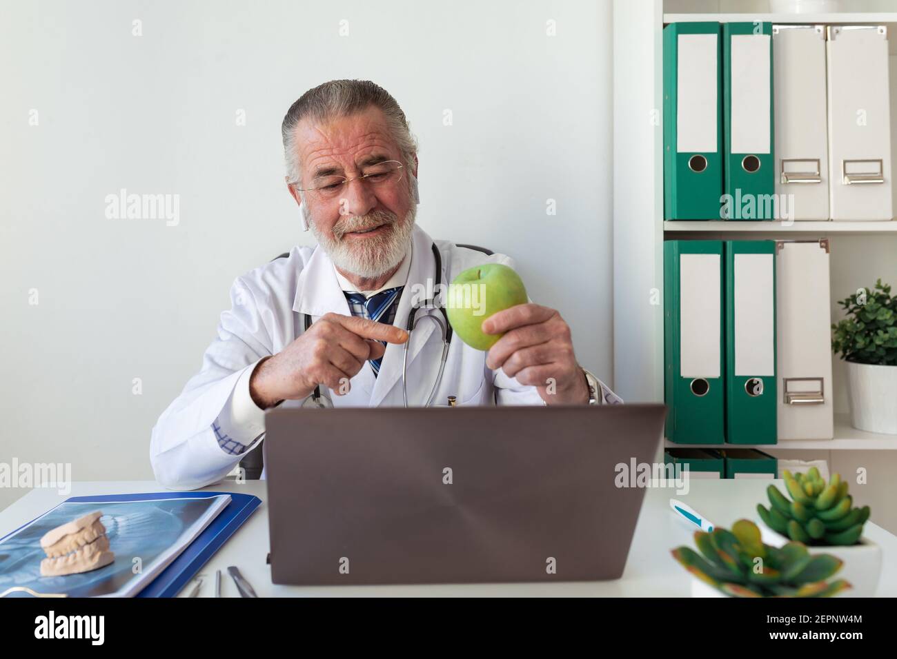 Elderly male orthodontist showing green apple against portable computer while taking on video call in clinic Stock Photo