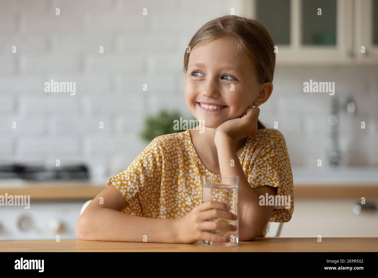 Healthy small girl dreaming by table holding glass of water Stock Photo