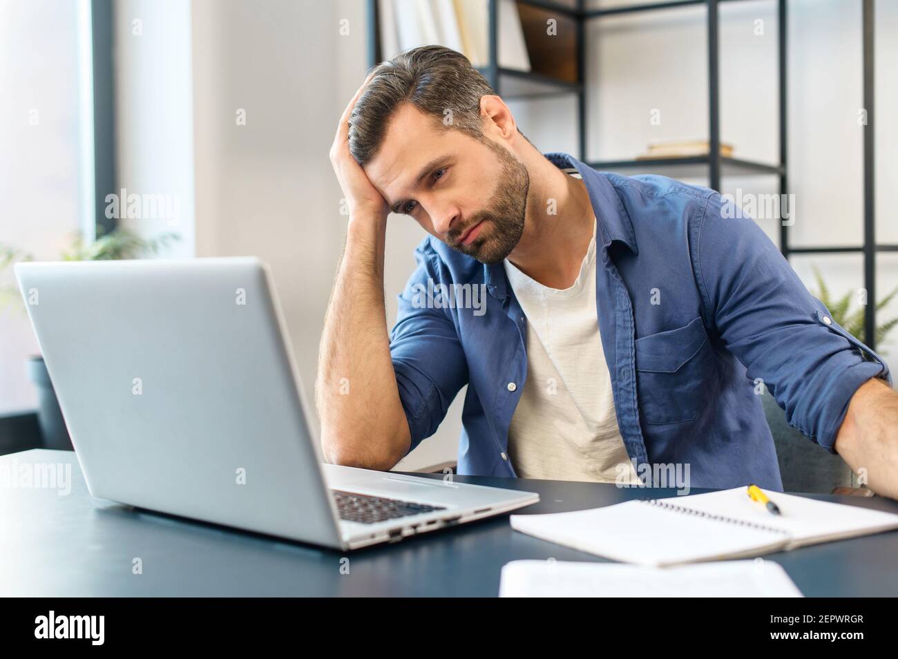 Young tired bearded office worker freelancer in casual clothes sitting at the desk at his workplace, supporting head with hand and looking at the laptop screen, feeling overworked and sleepy Stock Photo