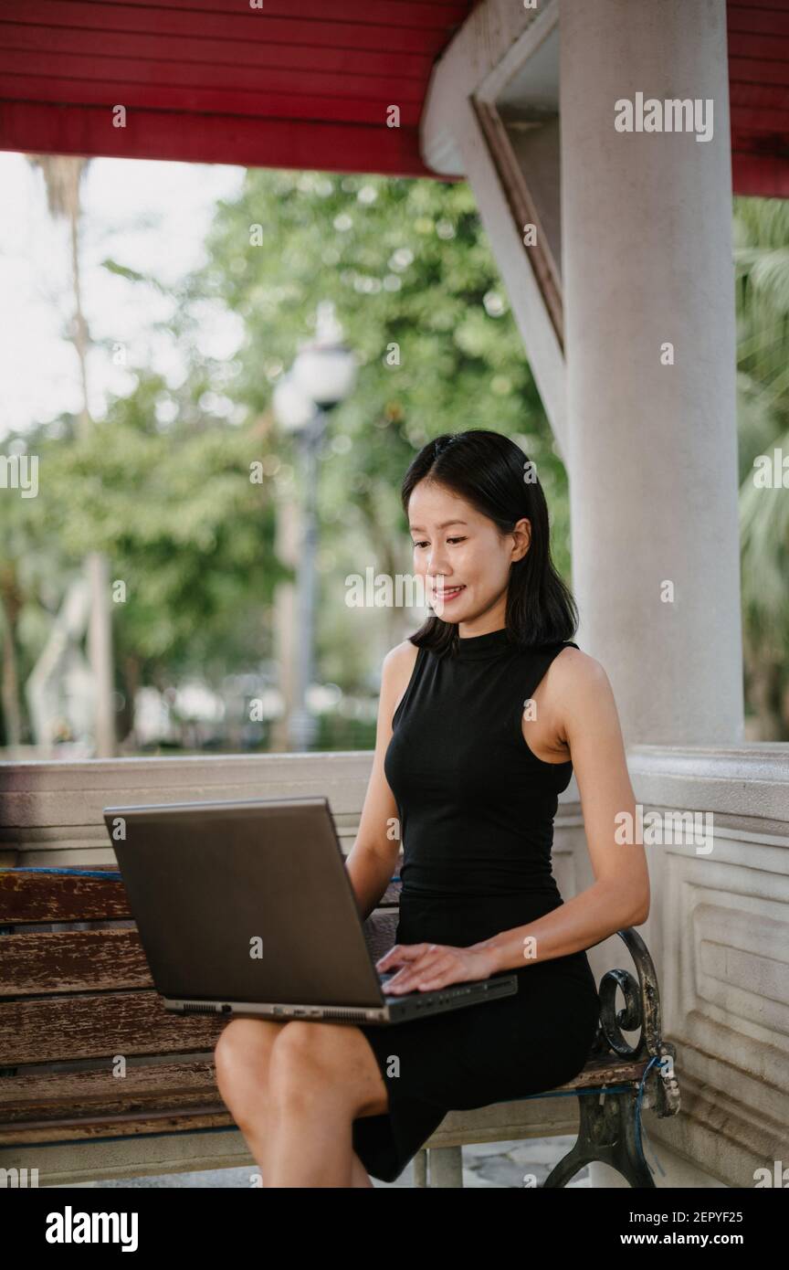 An attractive Asian businesswoman is sitting on a bench in a city park, working on her laptop, smiling. Stock Photo
