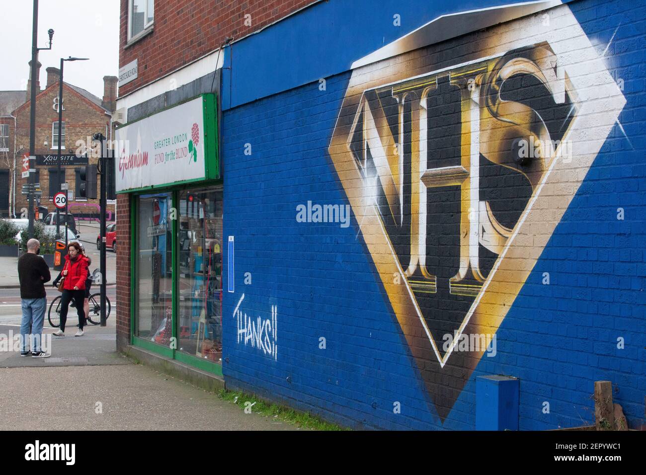 London, UK, 28 February 2021: A mural in Tulse Hill depicts the NHS logo in the style of a superhero badge. Passers-by take exercise or queue for the Co-op store round the corner, some wearing face masks and all socially distancing. Anna Watson/Alamy Live News Stock Photo