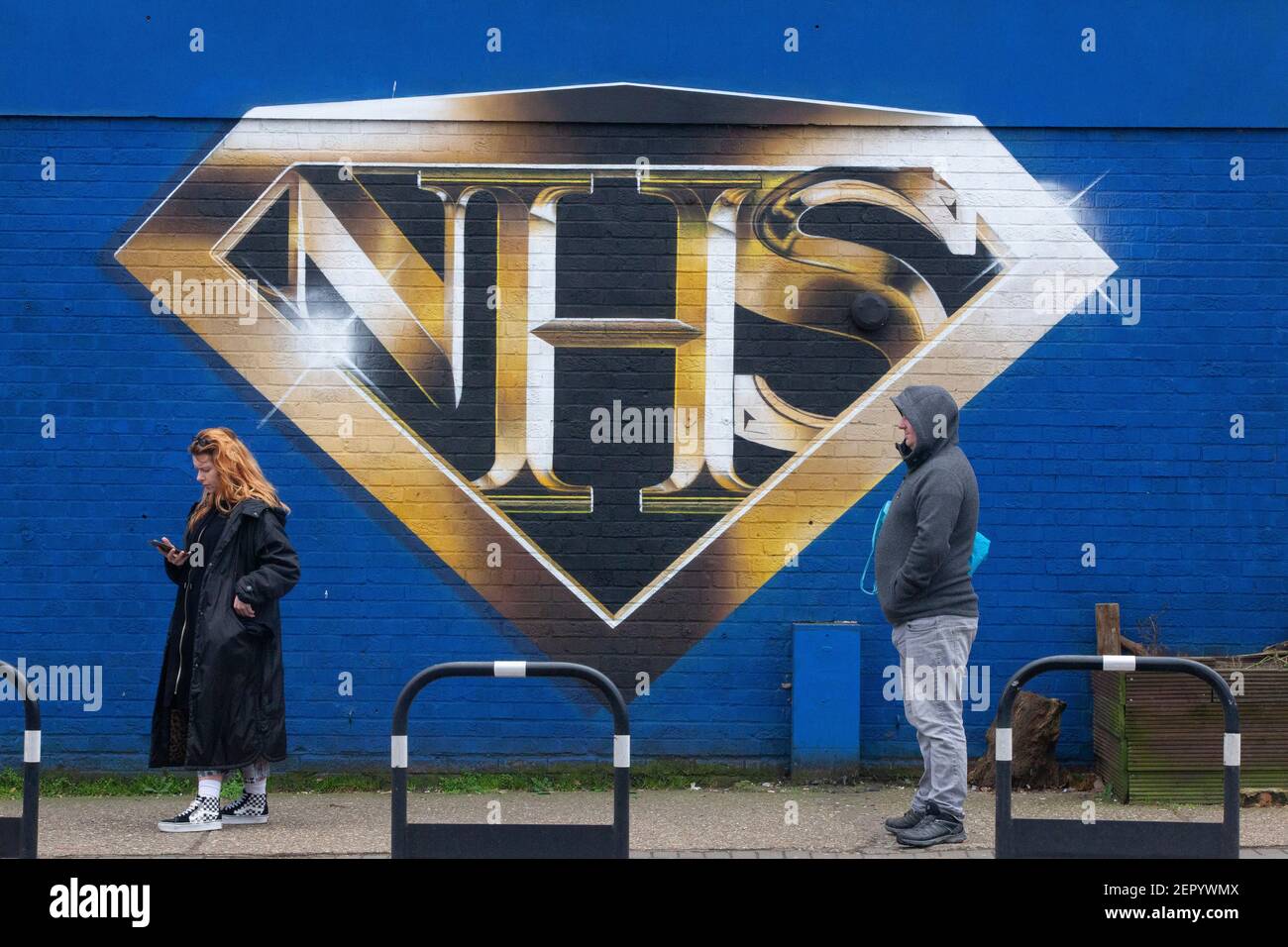 London, UK, 28 February 2021: A mural in Tulse Hill depicts the NHS logo in the style of a superhero badge. Passers-by take exercise or queue for the Co-op store round the corner, some wearing face masks and all socially distancing. Anna Watson/Alamy Live News Stock Photo