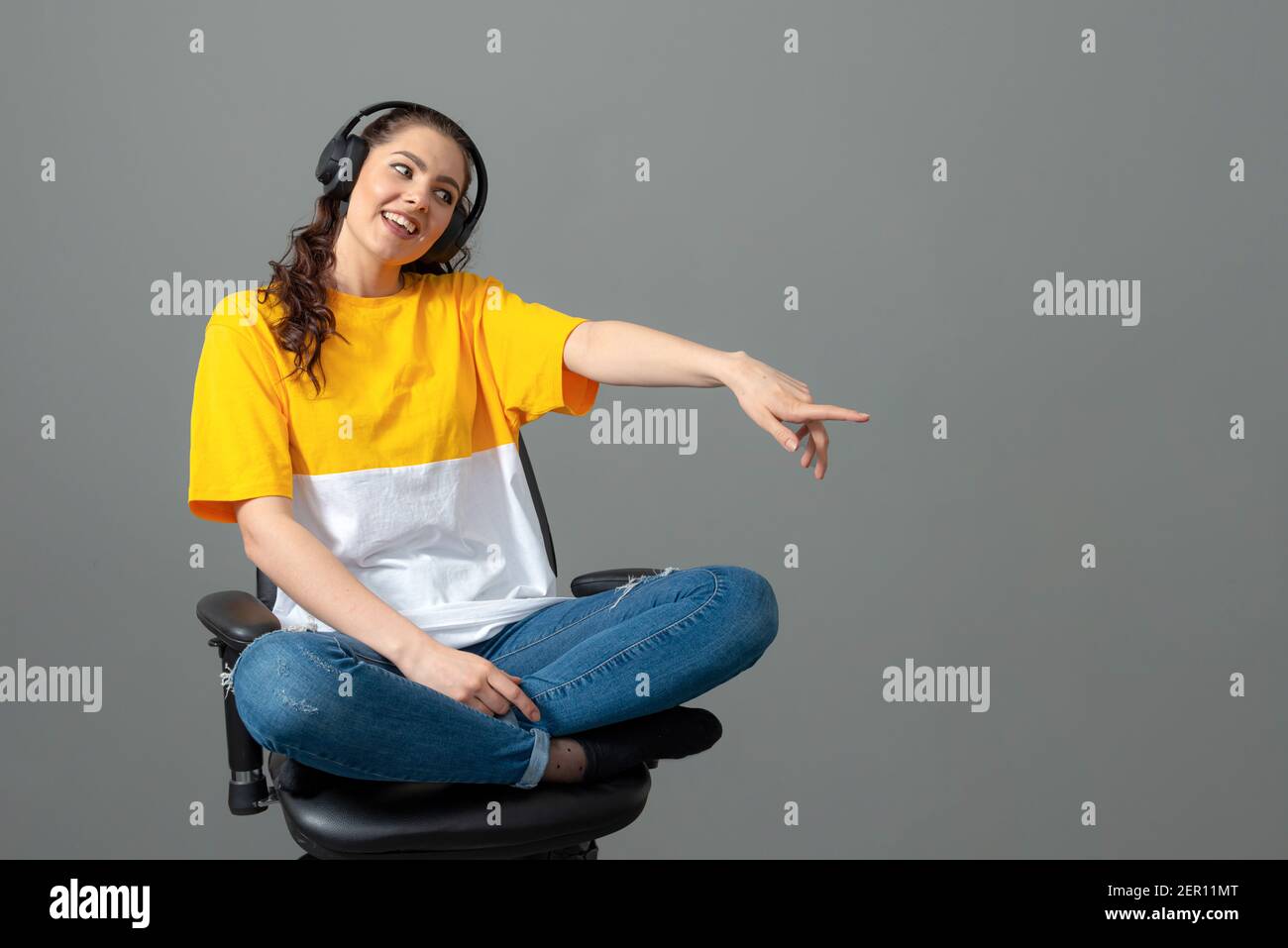 teenager with long wavy hair dressed in a yellow t-shirt sitting on an office chair and listening music, play games, listen to music, isolated on gray Stock Photo