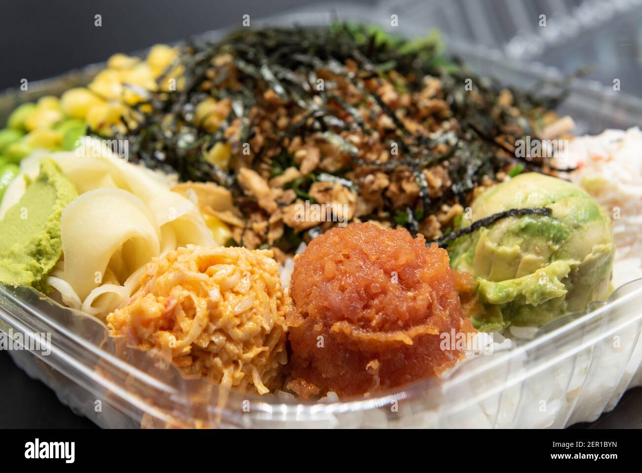 Large takeout plastic bowl with three round scoops of poke topped with shrimp tempura, crab meat, edemame, and corn served up and ready to eat. Stock Photo