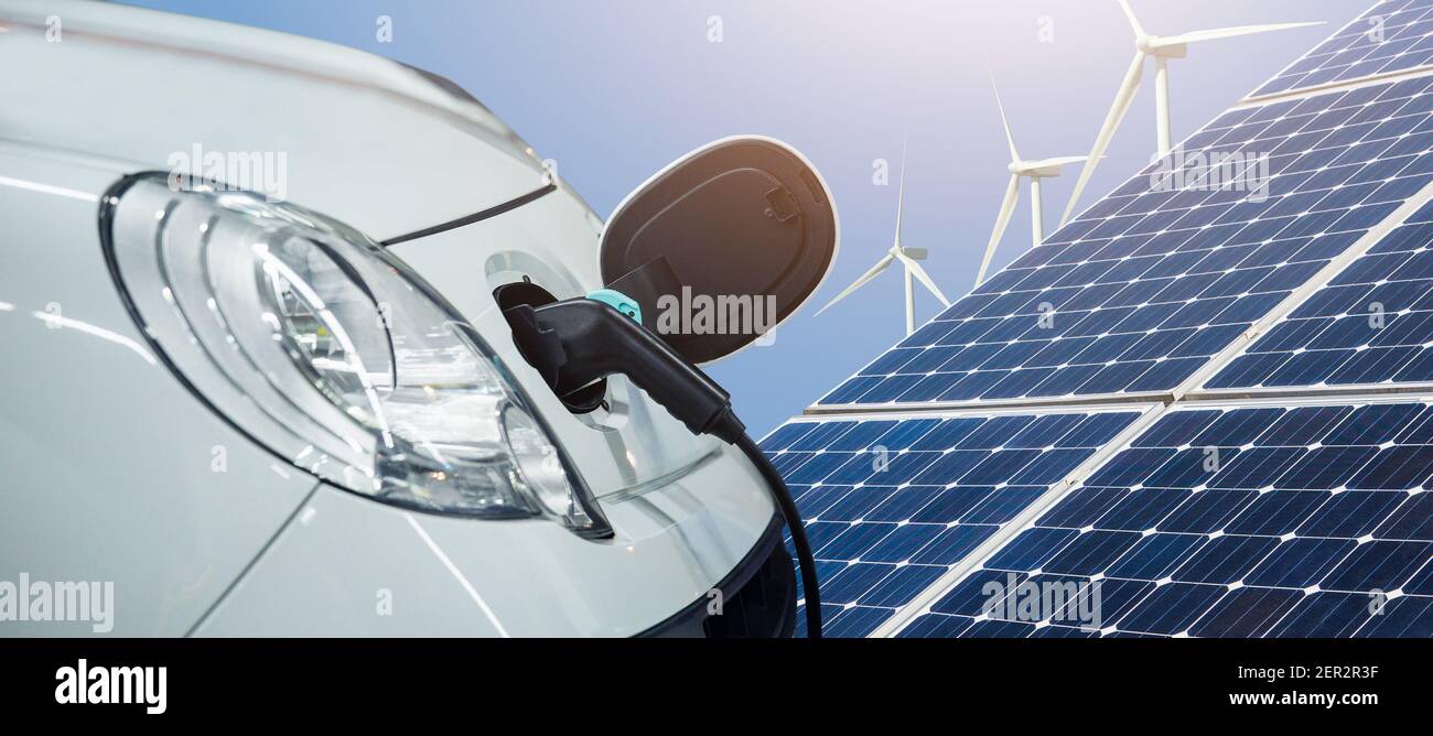 Close up of electric car with a connected charging cable on the background of solar panels and wind turbines - sources of clean renewable energy Stock Photo