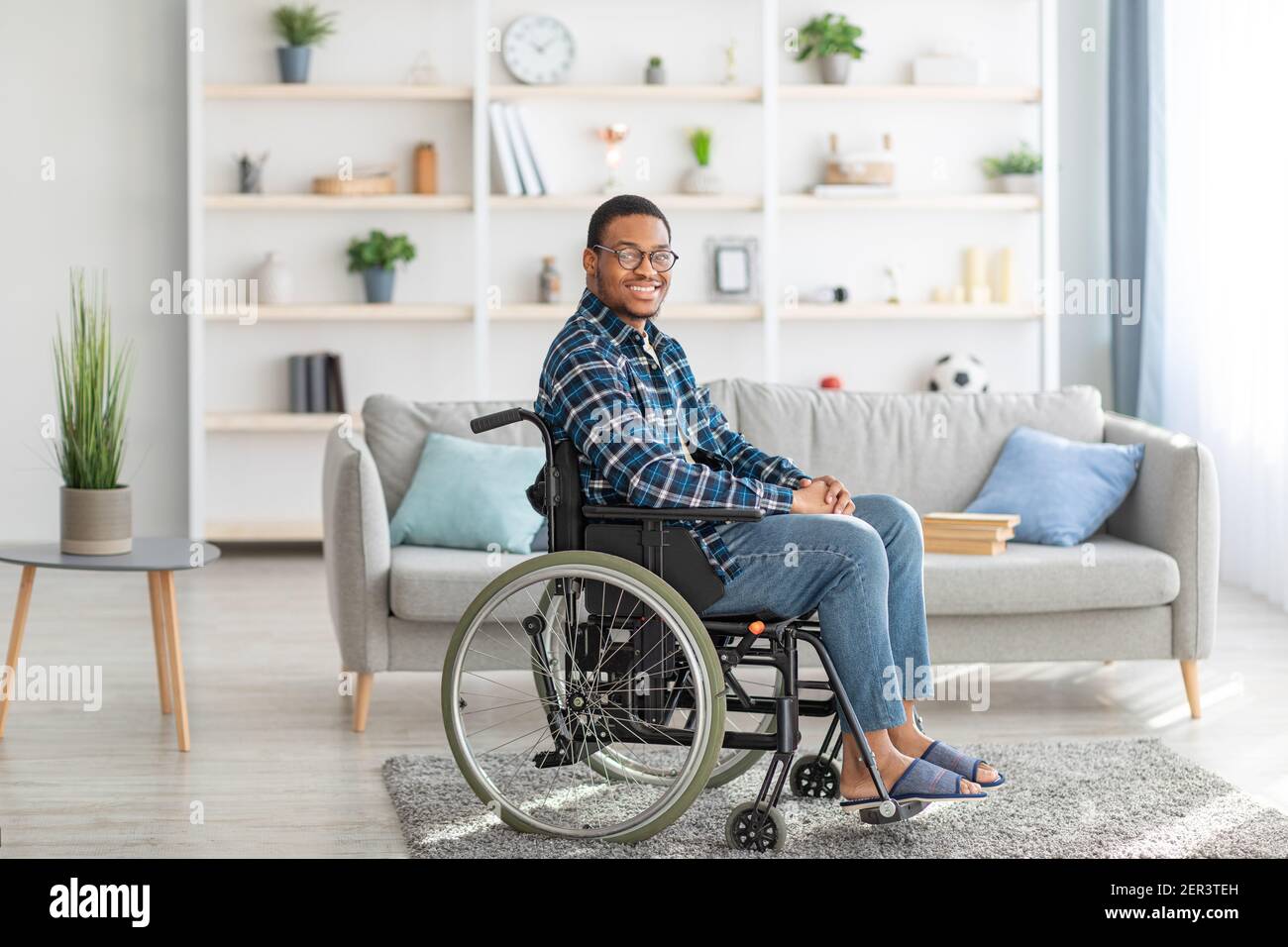 Cheerful handicapped black guy in wheelchair smiling and looking at camera in living room Stock Photo