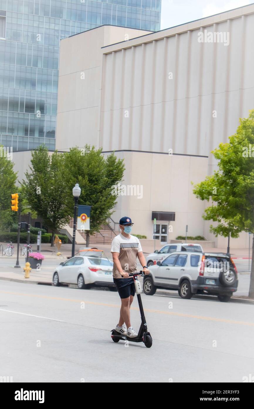 07 25 2020 Tulsa USA Young man wearing shorts and teeshirt and ball cap and face mask  ridess electric scooter down city street with large buildings i Stock Photo