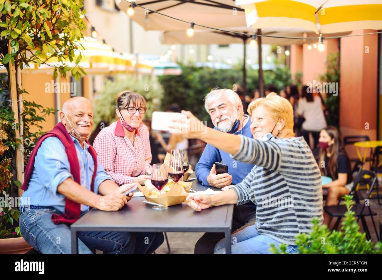 Senior couples drink red wine at bar with face masks protection - New normal lifestyle concept with happy people having fun together taking a selfie Stock Photo