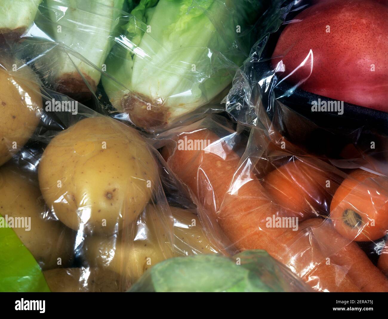 Unnecessary packaging - clear plastic bag and cellophane wrapping on vegetables sold in a supermarket (including potatoes, carrots and lettuce). Stock Photo