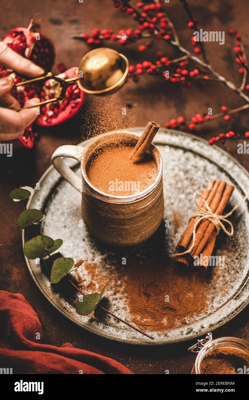 Turkish traditional wintertime hot drink Salep. Human hand pouring cinnamon powder to sweet warming Salep drink over rusty table background. Turkish s Stock Photo