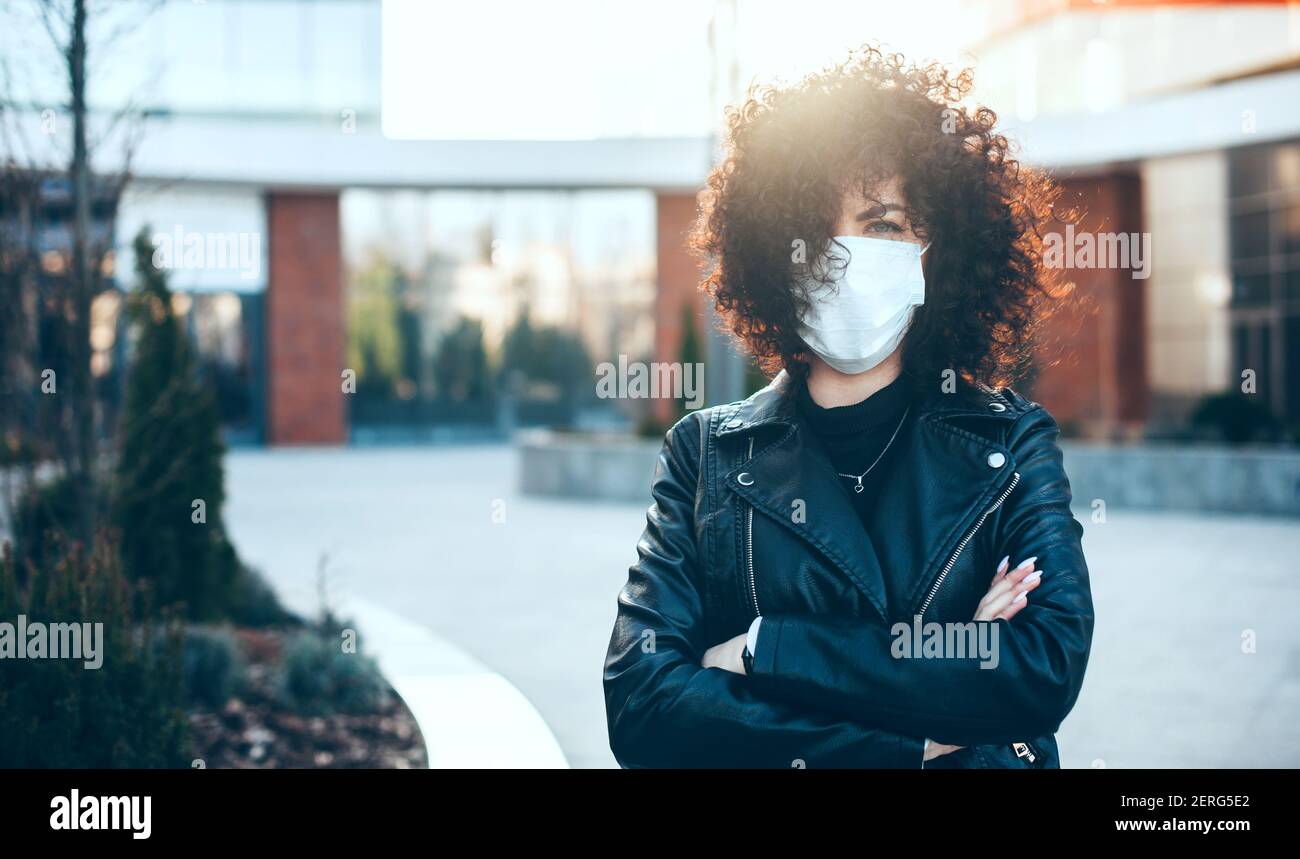 Caucasian woman with crossed hands and curly hair posing outside with a medical mask looking at camera Stock Photo