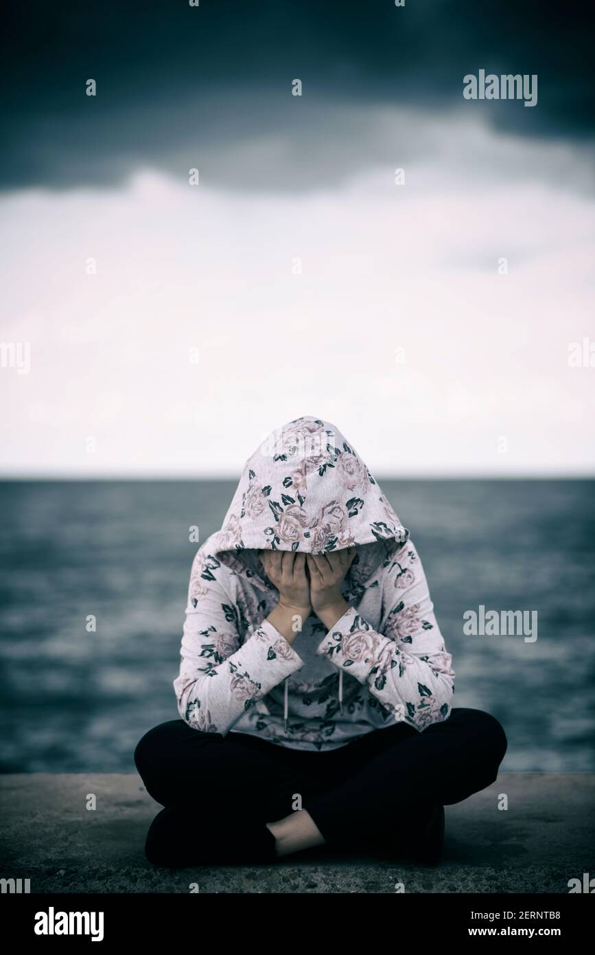Miserable hooded woman sitting on the beach hiding face with hands crying Stock Photo