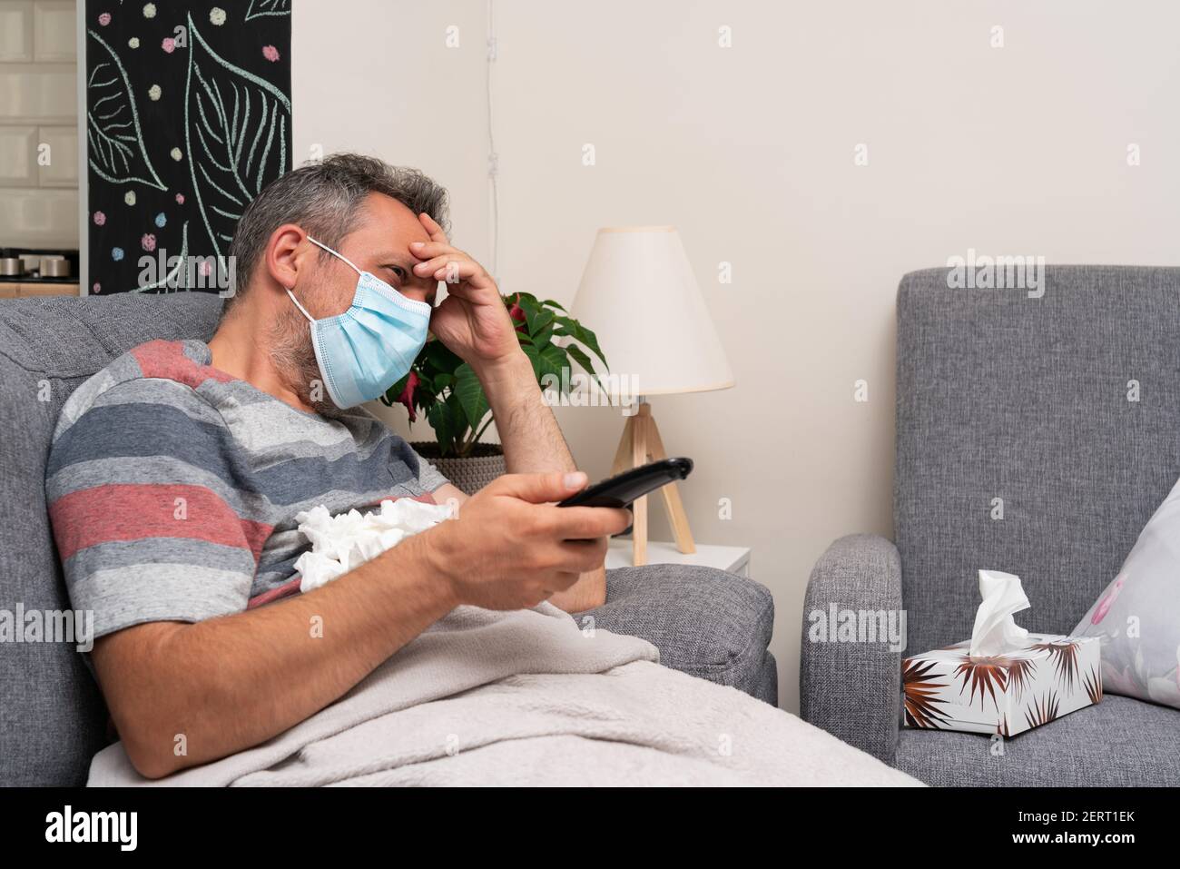 Sick adult man having migraine or high fever sitting on home sofa holding tv remote control zapping wearing surgical or medical mask to prevent sars c Stock Photo