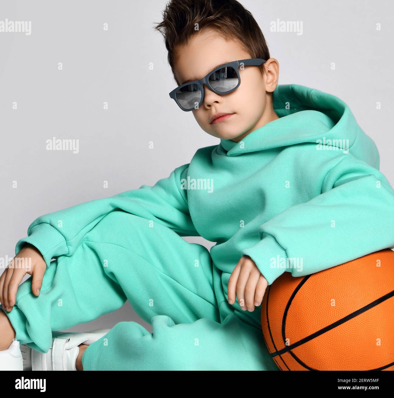 Portrait of cool frolic handsome kid boy, school gang leader in modern sunglasses and modern green, mint color sportswear hoodie and pants sitting wit Stock Photo