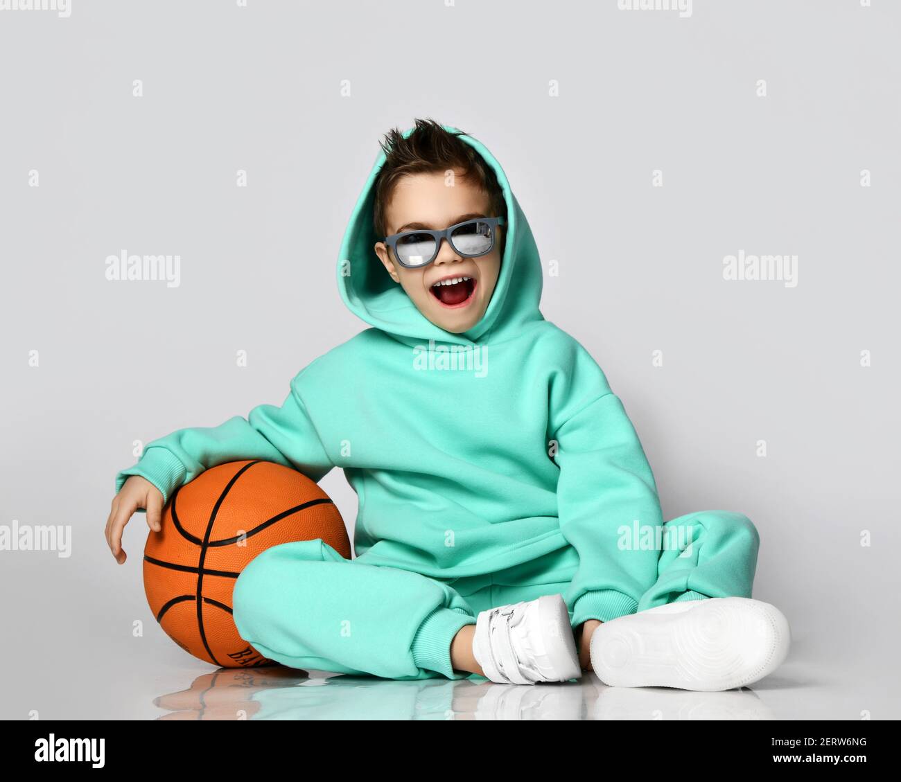 Happy screaming laughing frolic active kid boy in modern green, mint color sportswear hoodie, pants and sneakers sitting with basketball and his hood Stock Photo