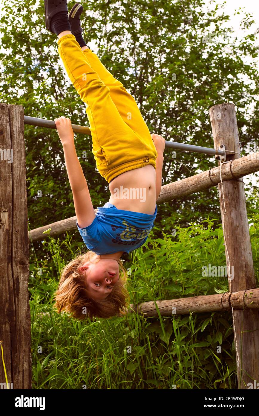 A boy in yellow pants makes a stand on the horizontal bar upside down. Sports activities in nature. Active childhood. Stock Photo