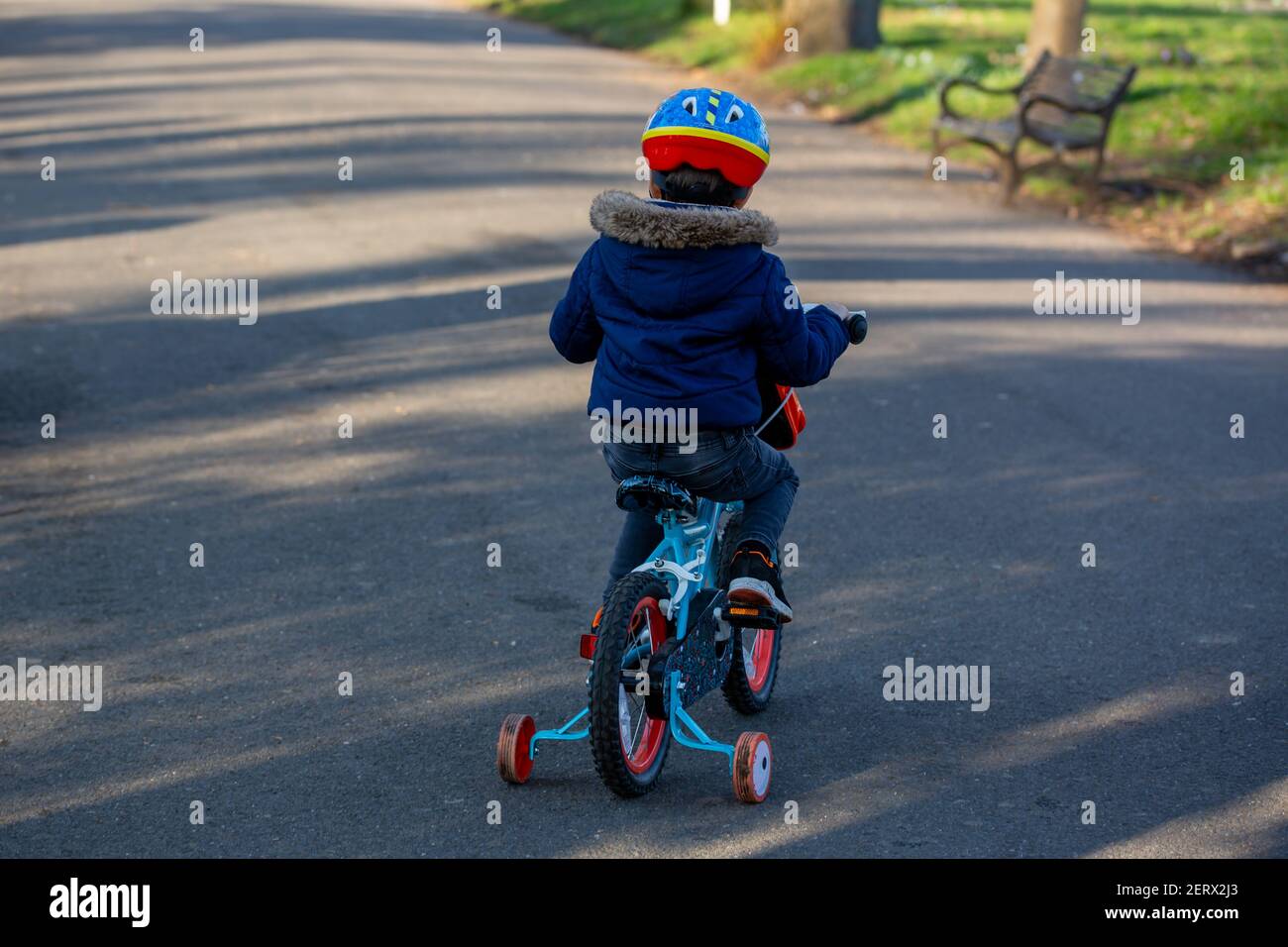 A small boy wearing a cycle helmet riding a bike in the park with stabilisers or stabilizers Stock Photo