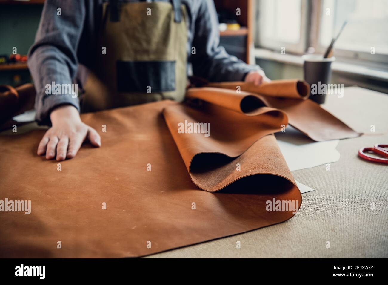 A young apprentice in a boot workshop prepares leather for further use on a large table Stock Photo