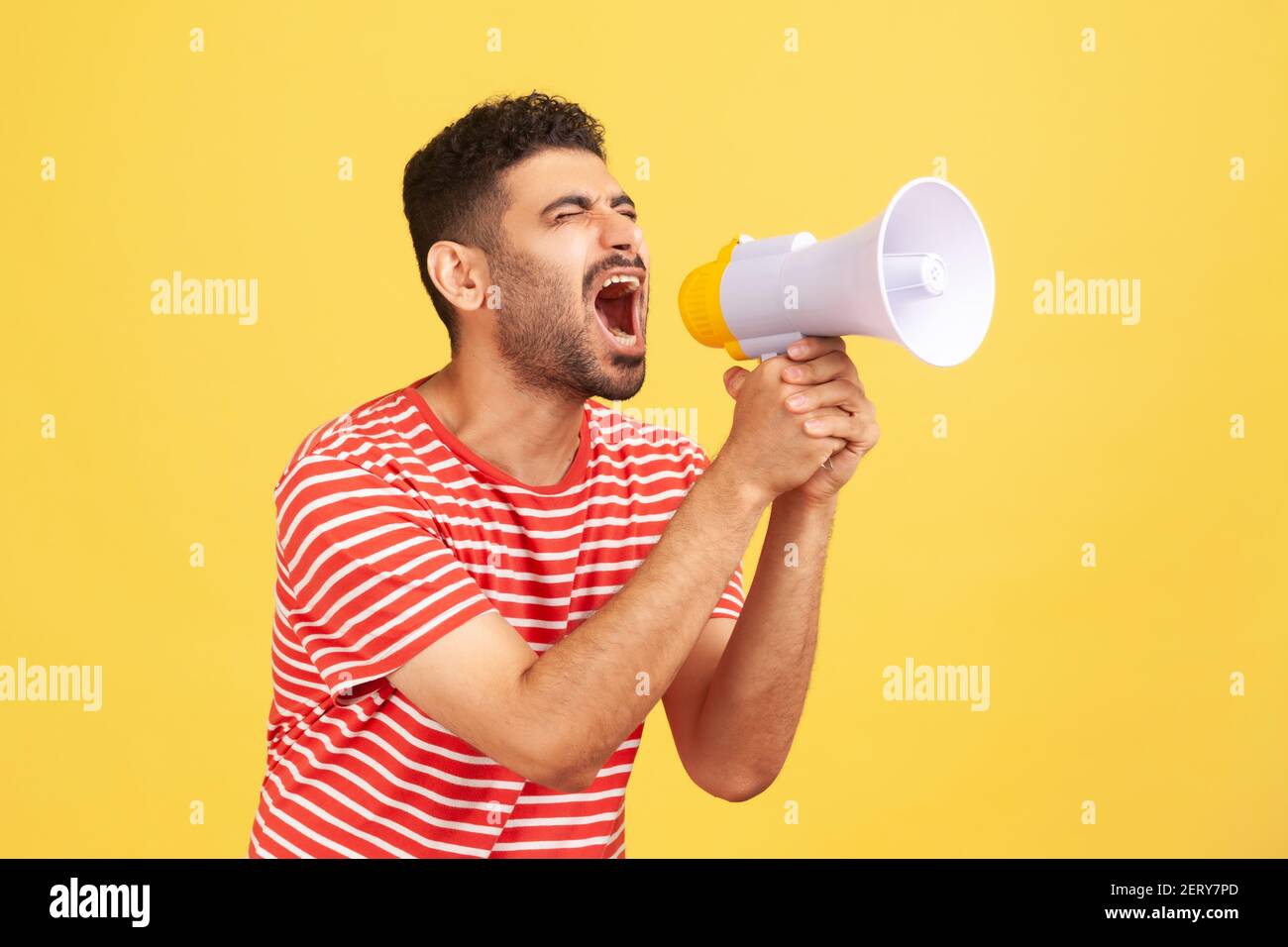 Angry nervous man with beard in striped t-shirt loudly screaming at megaphone, making announce, protesting, wants to be heard. Indoor studio shot isol Stock Photo