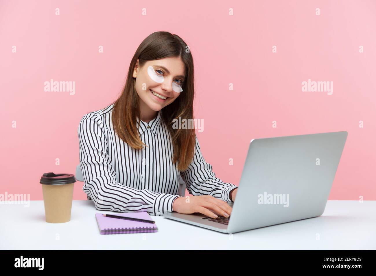 Smiling brunette business woman in striped shirt working on laptop sitting at home office with eye patches, good mood. Indoor studio shot isolated on Stock Photo
