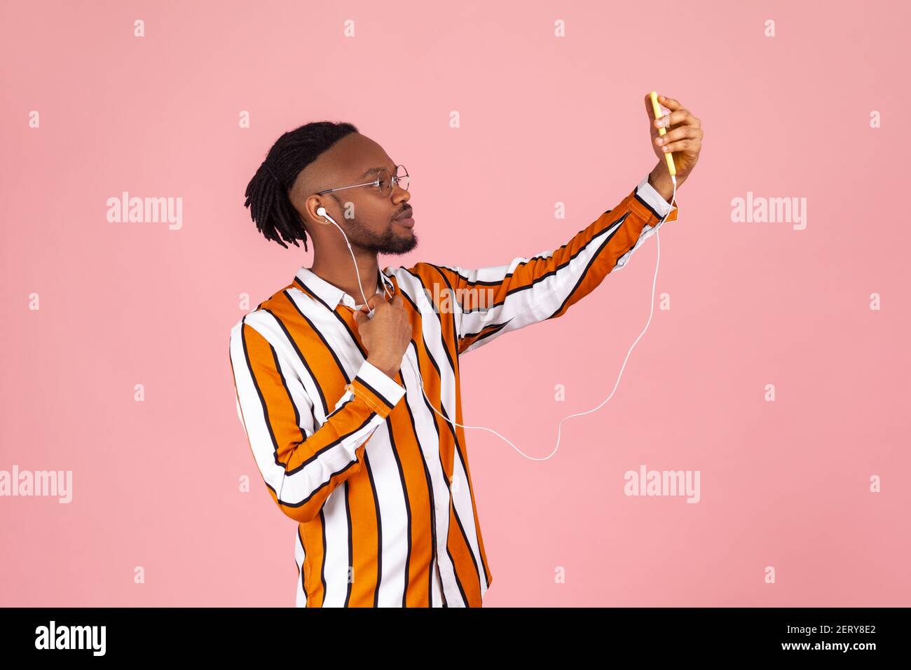Side view african man with dreadlocks and ear piercing in striped shirt making selfie on smartphone frontal camera using wire earphones. Indoor studio Stock Photo