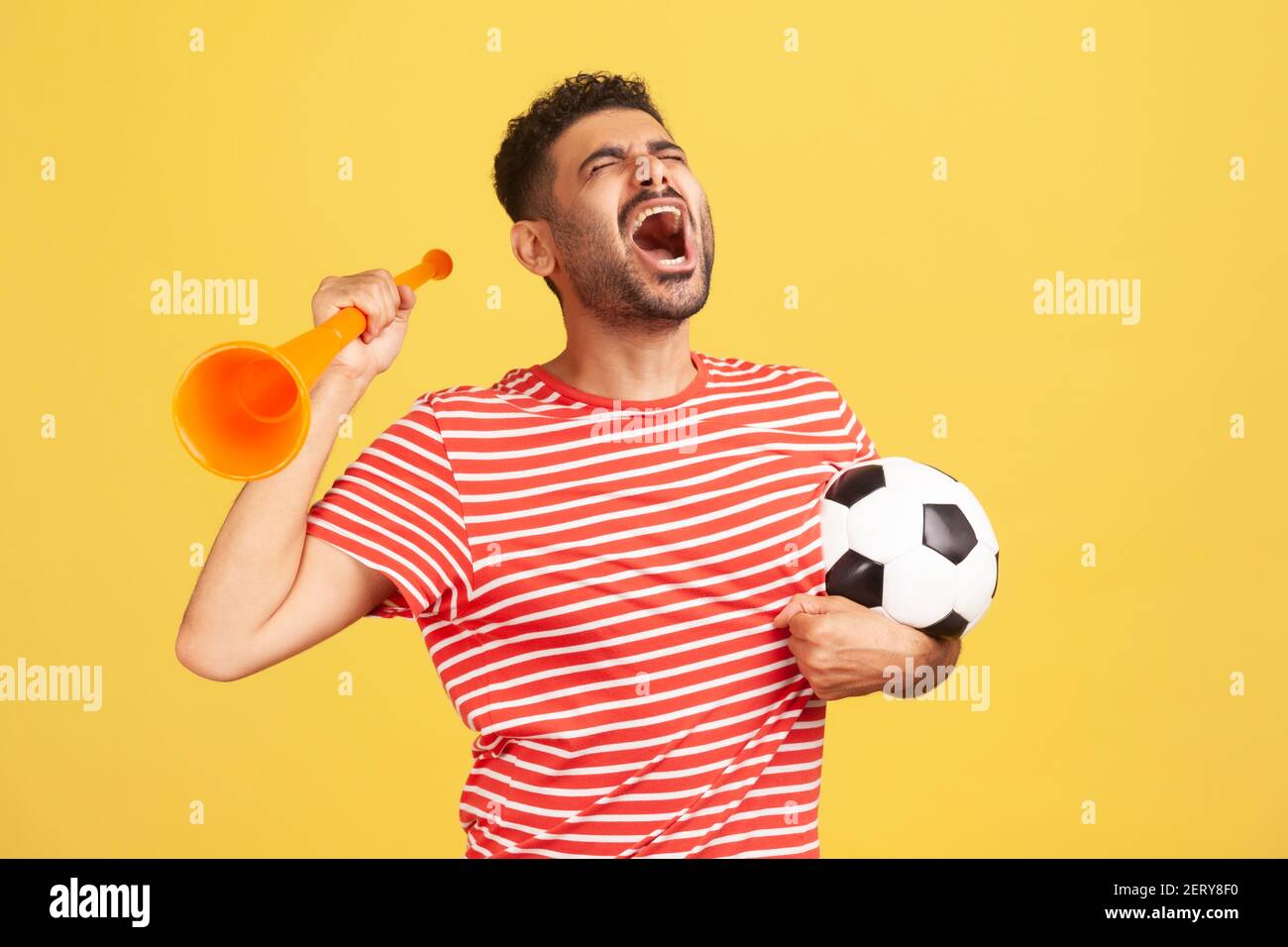 Bearded man in striped t-shirt screaming widely opening mouth, celebrating victory of favourite football team, holding horn and ball in hands. Indoor Stock Photo