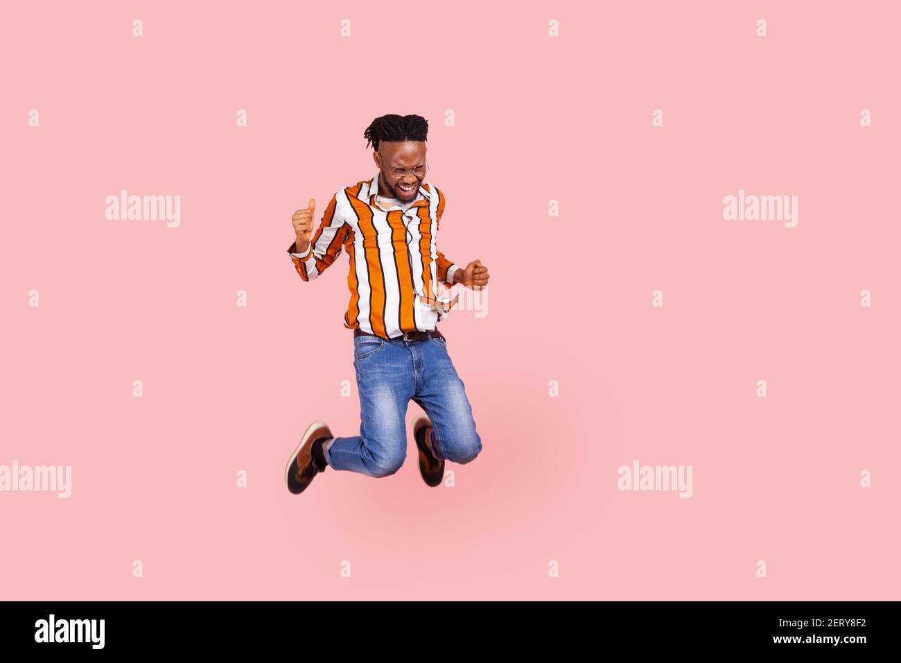 Full length happy positive african man with dreadlocks in stylish bright shirt highly jumping on trampoline, having fun, enjoying like in childhood. I Stock Photo