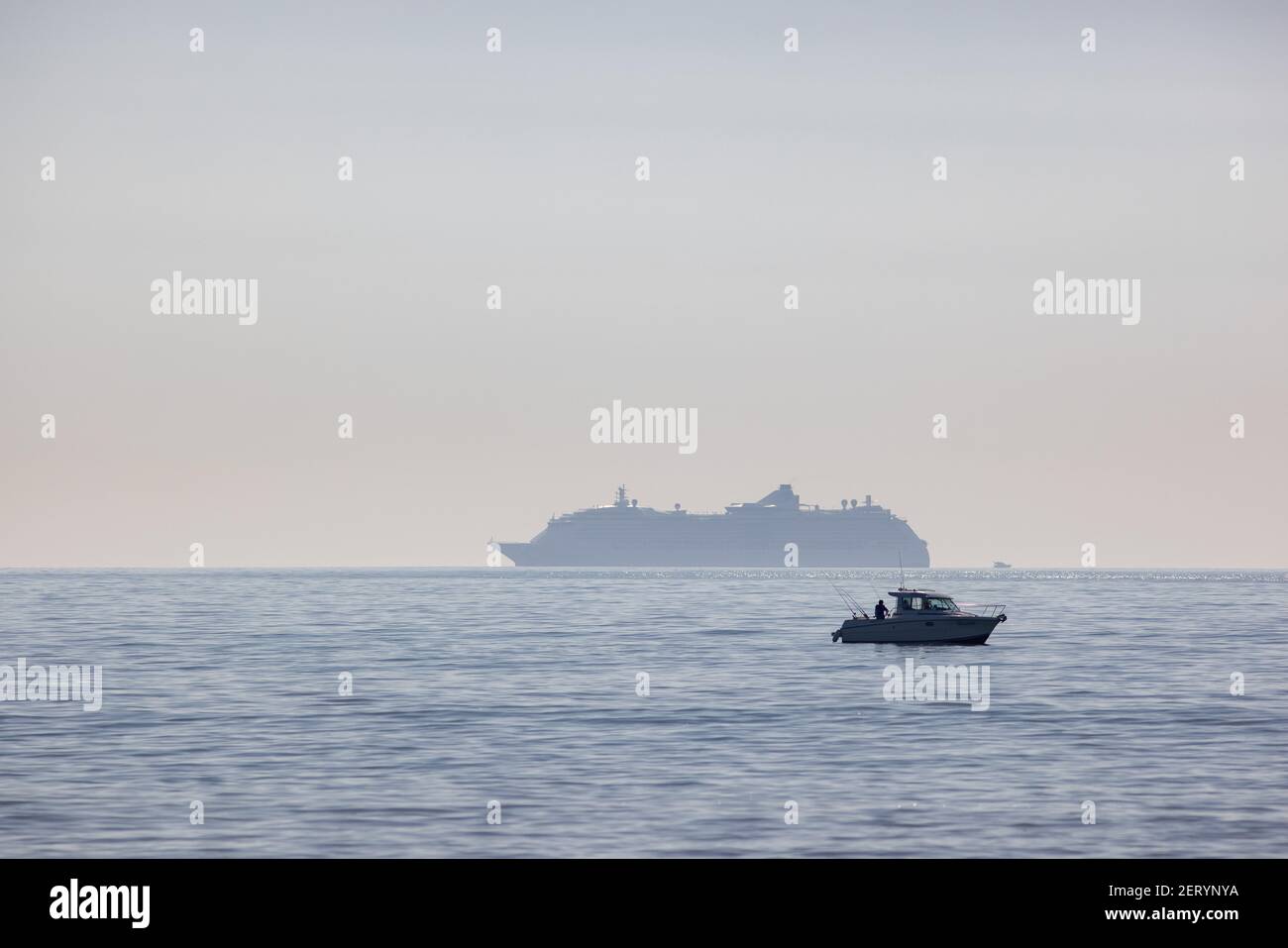 A fishing boat just off of the shore with rods in action as a cruise ship sits in the mist near the beach at Fisherman’s Walk, Southbourne, Bournemout Stock Photo