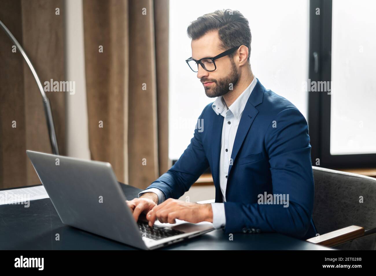 Serious concentrated determined young bearded office worker in business casual clothes, sitting at the desk, typing on laptop, businessman professiona Stock Photo
