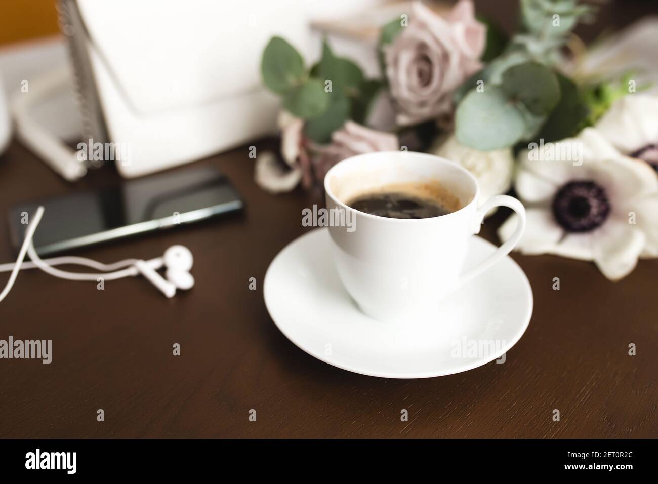 A cup of coffee with a bouquet of tender anemone, eucalyptus branches and pastel purple roses, a woman handbag, headphones and a phone on a brown wood Stock Photo
