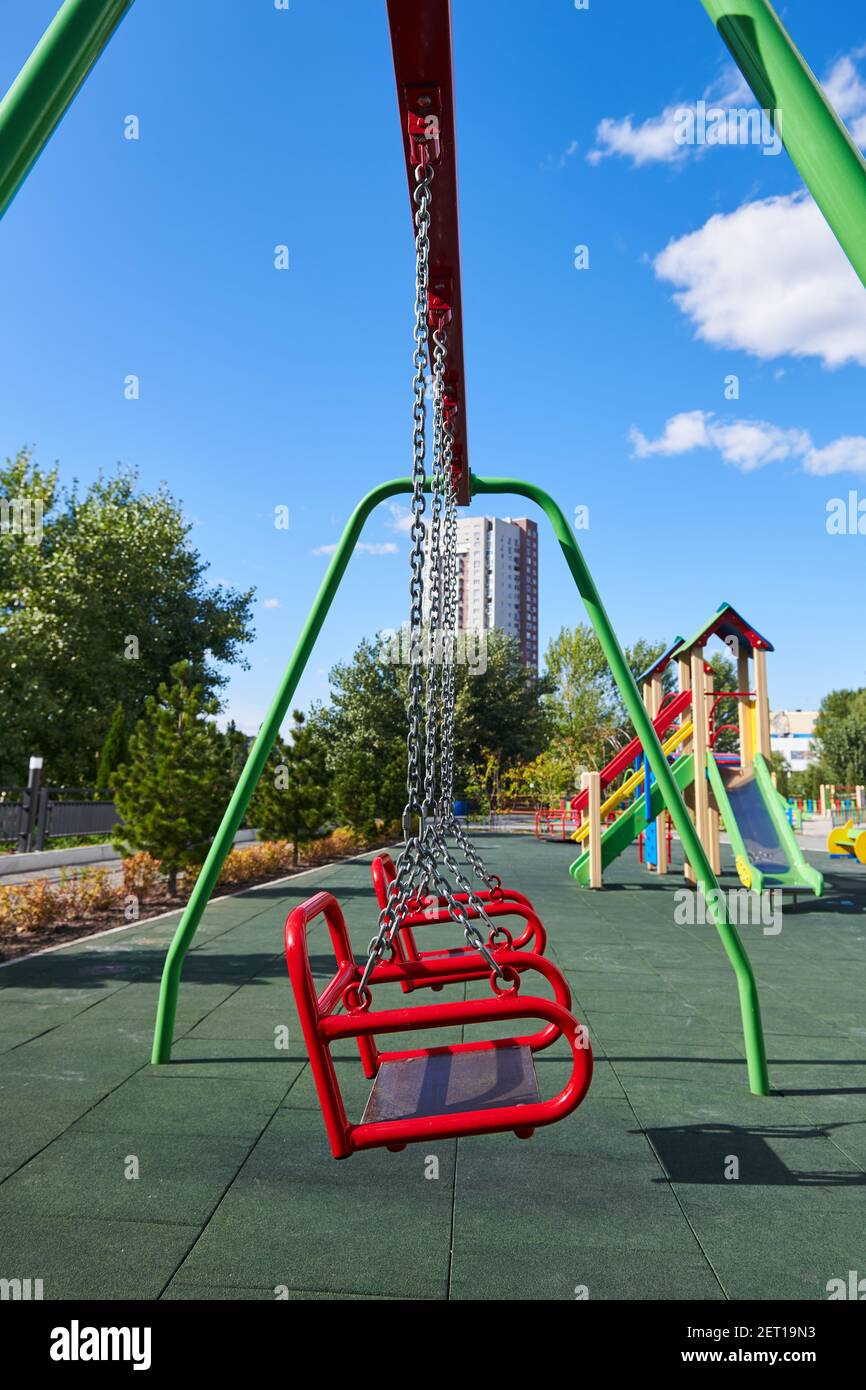 Empty red swings on the playground against blue sky and trees. Swing is hanging on the metal chains. Empty place during coronavirus pandemic. Covid-19 Stock Photo