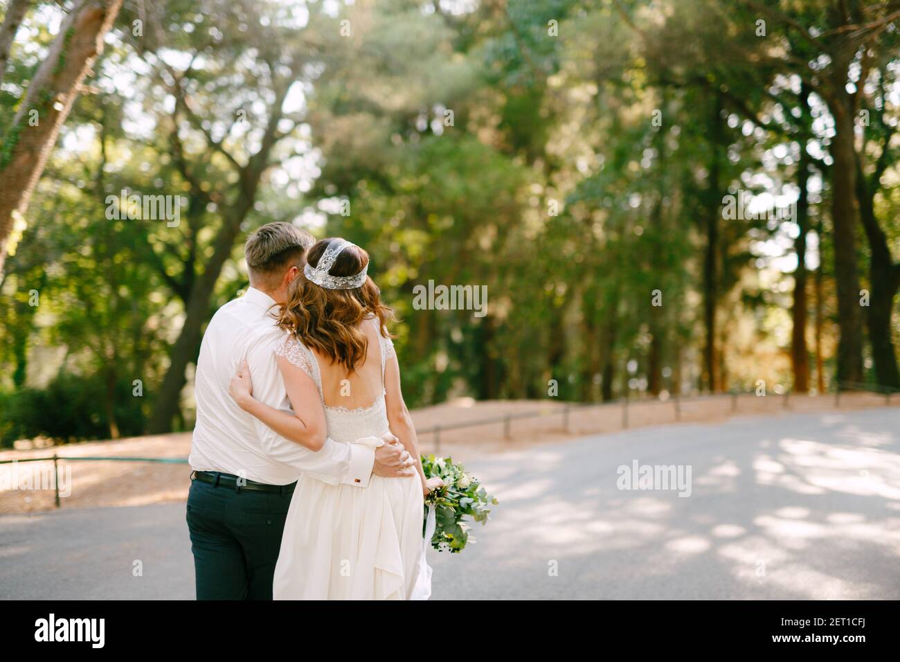The bride and groom are walking along the road in the park hugging each ...