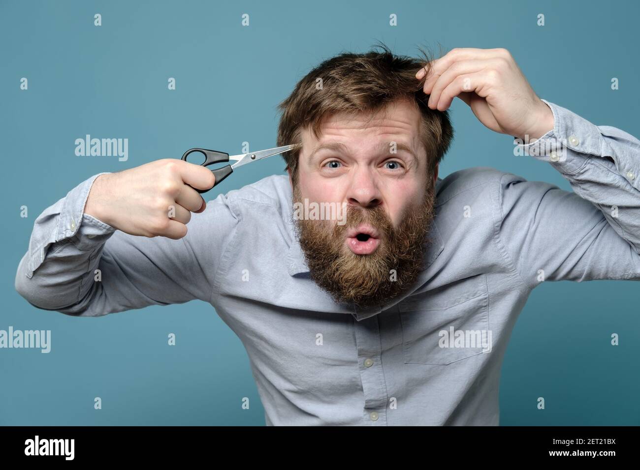 An annoyed, shaggy man holding hair scissors to his head as if it were a gun, he had not visited a barbershop for a long time. Stock Photo
