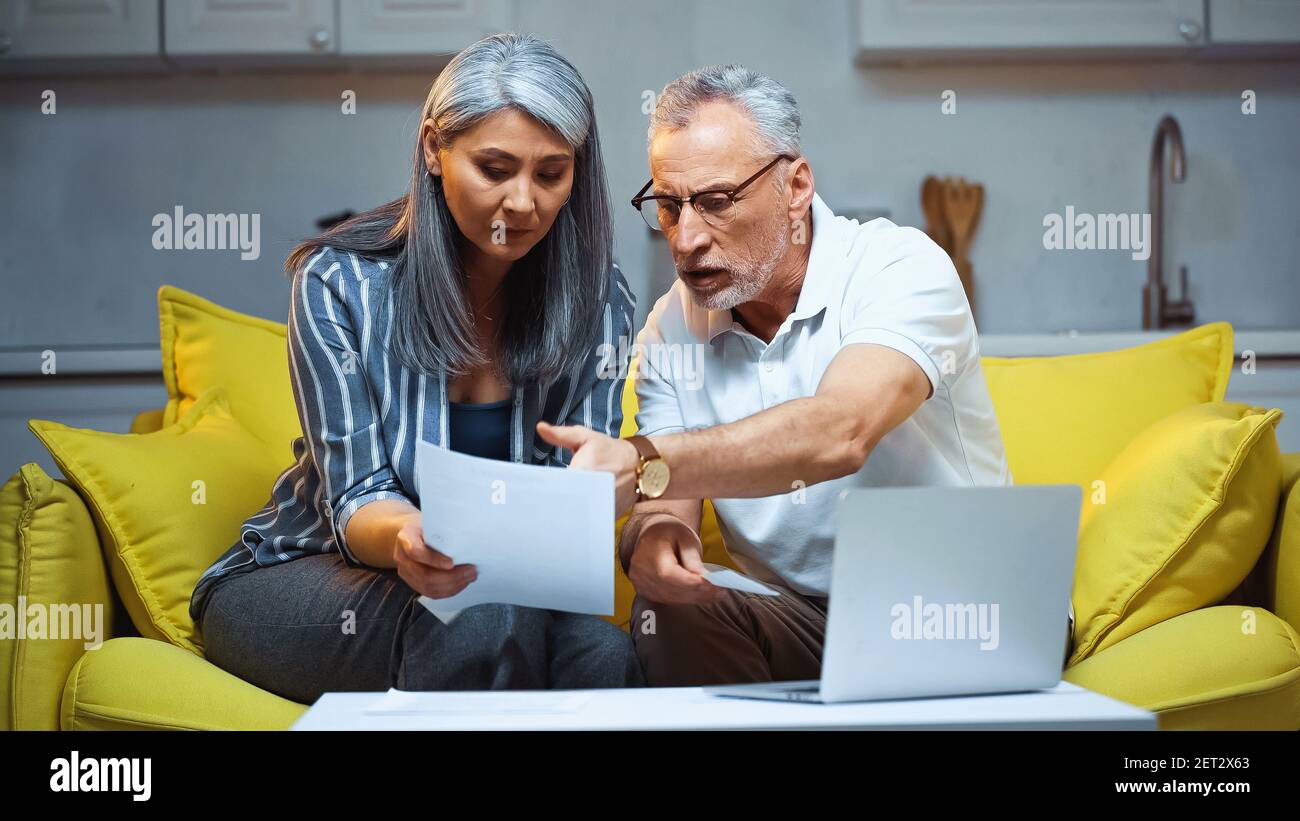 serious man pointing with hand at document near laptop and asian wife Stock Photo
