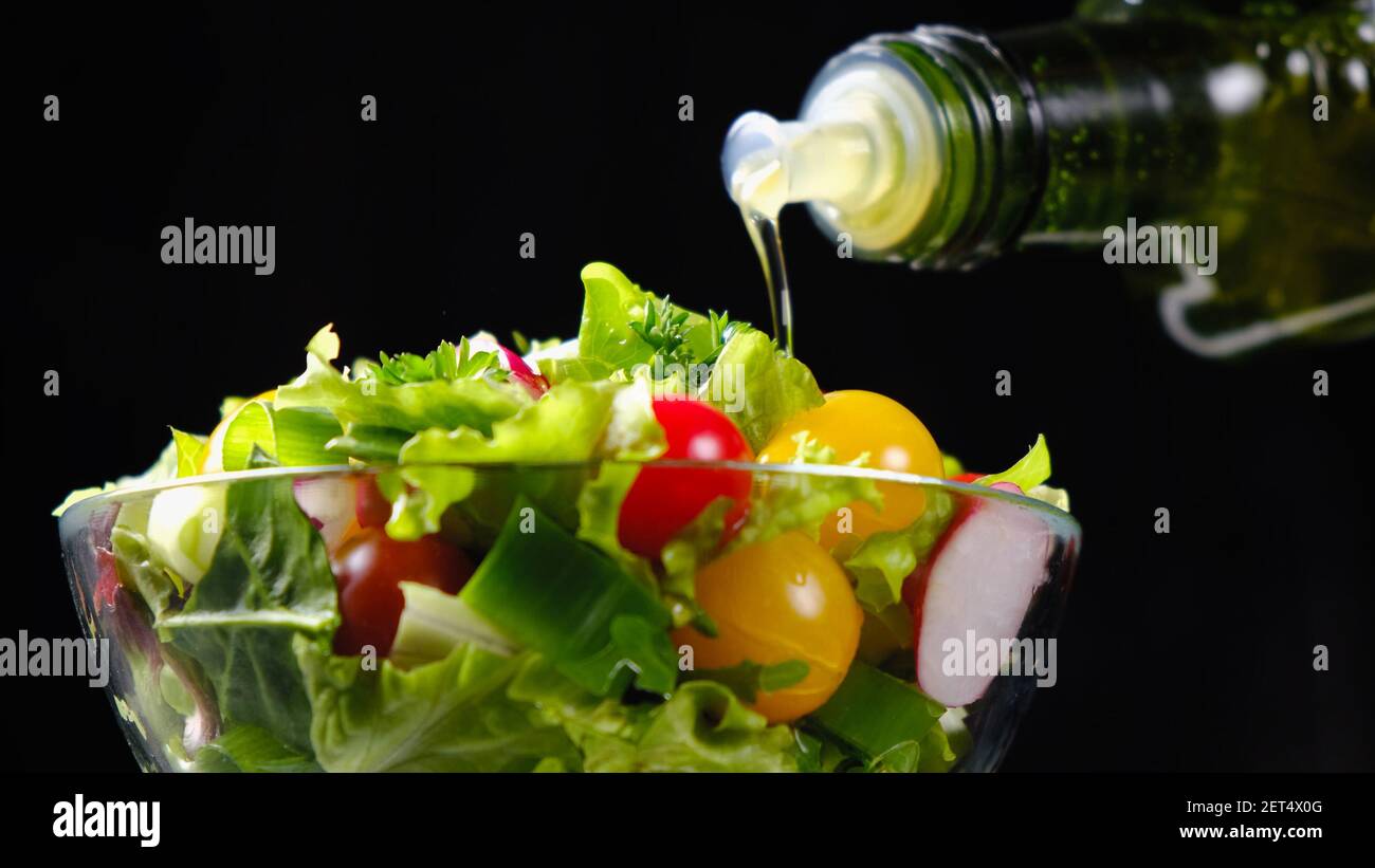 Olive oil pouring from bottle onto vegetable salad over black background Stock Photo