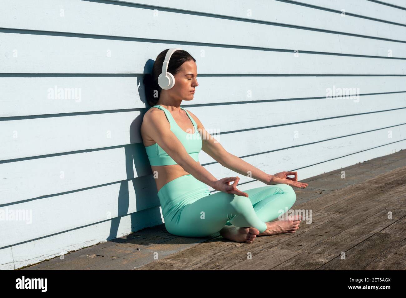 Woman meditating practicing yoga sitting in the sun wearing headphones with a blue clapboard background. Stock Photo