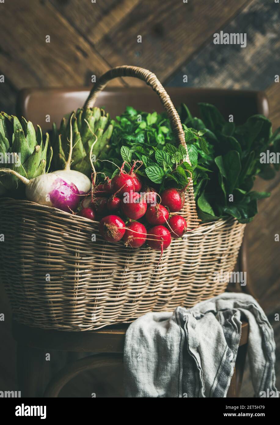 Basket of fresh organic garden vegetables and greens on chair, rustic wooden wall at background Stock Photo