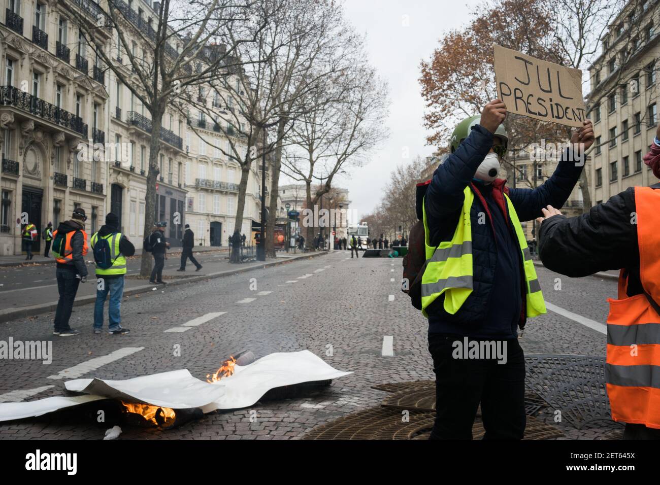 A demonstrator on Avenue Marceau holds a sign reading "JUL PRESIDENT,"  endorsing French rapper Jul for president as a fire burns in the street  beside them. The protests marked the fourth weekend