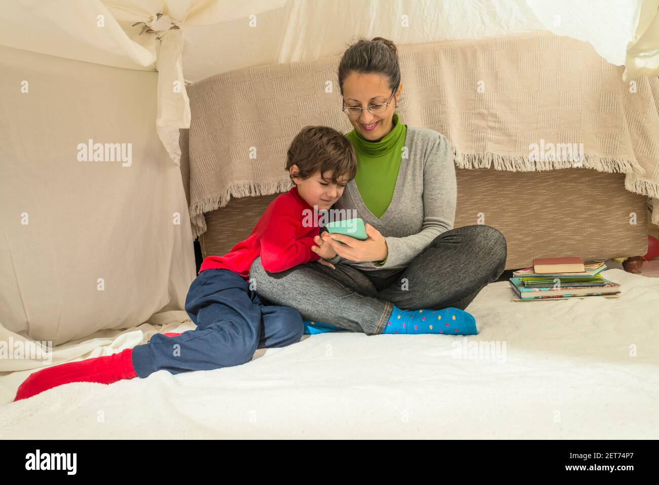 A woman and a child looking at a smart phone sitting on a blanket in a makeshift tent in the living room at home. Home lifestyle concept. Stock Photo